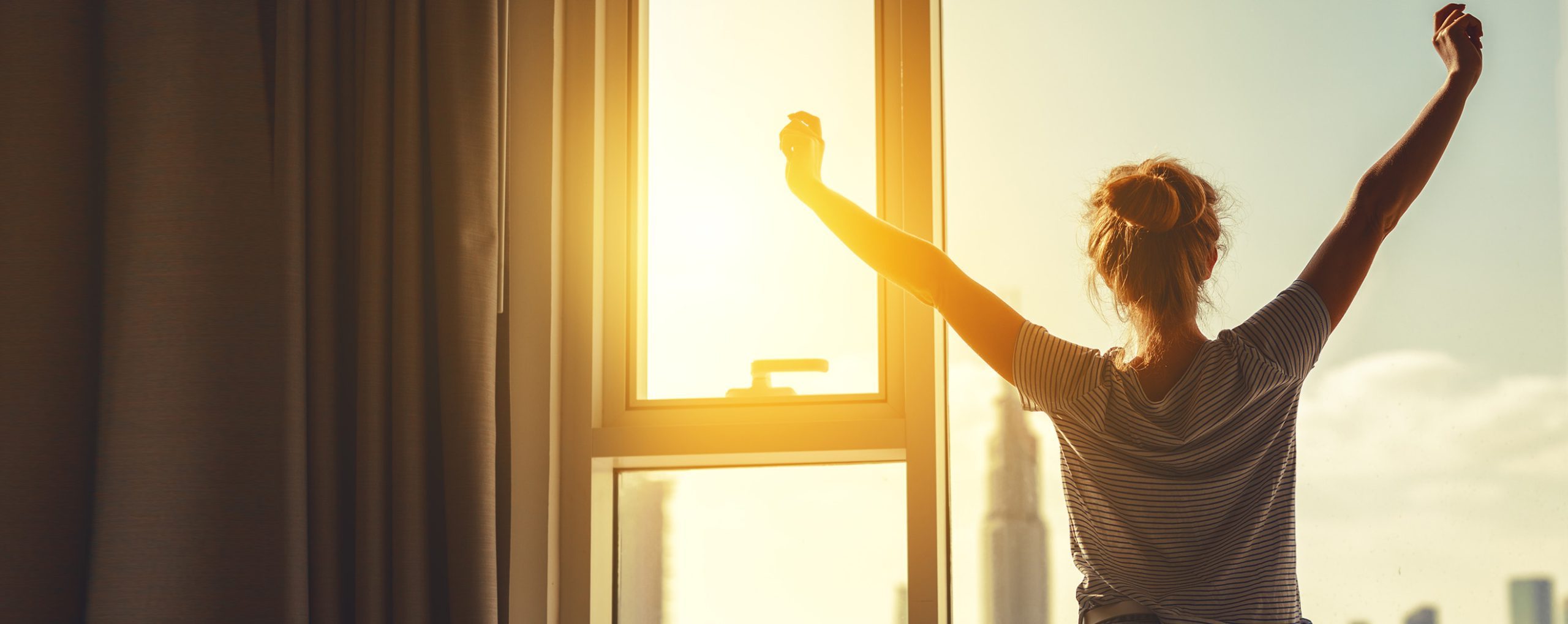 Frau streckt sich morgens vor einem Fenster, während die Sonne aufgeht.