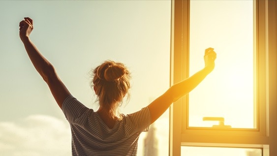 Frau streckt sich morgens vor einem Fenster, während die Sonne aufgeht.