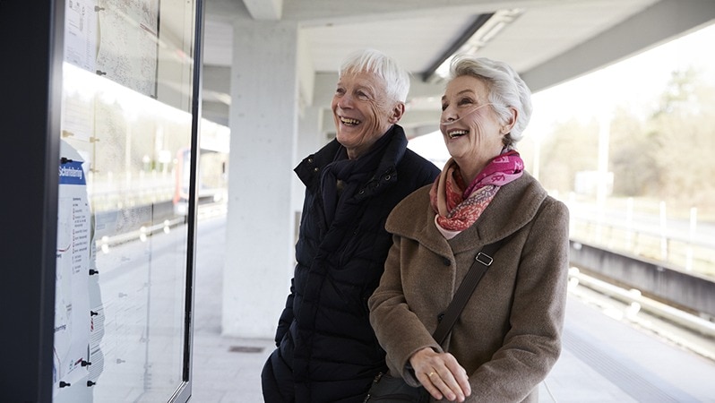 Ein älterer Mann und eine ältere Frau mit Sauerstoffkonzentrator lesen an einer Informationstafel auf einem Bahnsteig