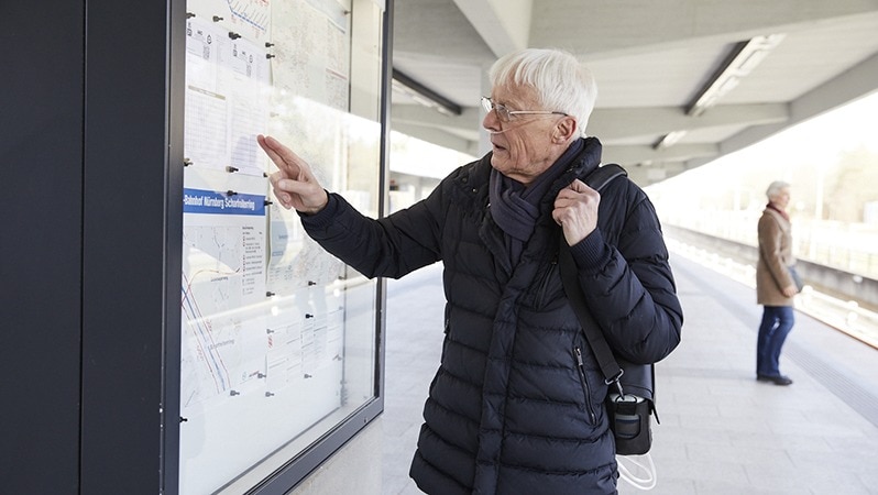 Älterer Mann mit Sauerstoffkonzentrator liest an einer Informationstafel auf einem Bahnsteig