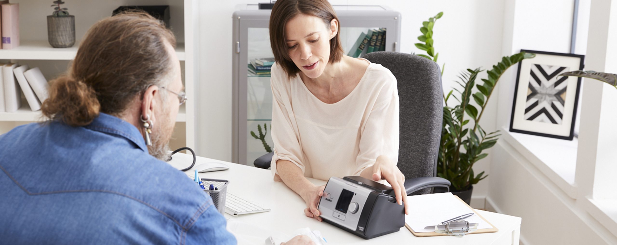 Eine Ärztin erklärt einem Patienten die Funktionsweise eines CPAP-Geräts während eines Beratungsgesprächs.