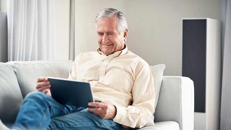 Ein älterer Mann sitzt entspannt auf einem Sofa und hält ein Tablet in der Hand, während er lächelt und den Bildschirm betrachtet.