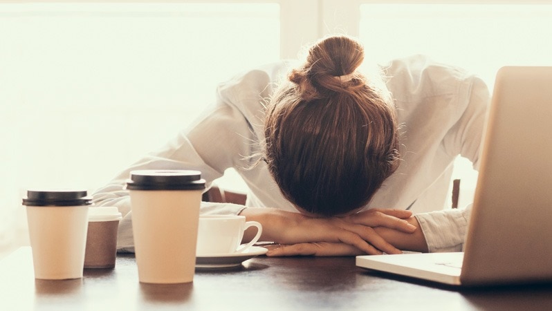 Frau liegt erschöpft mit dem Kopf auf dem Tisch neben mehreren Kaffeebechern und einem Laptop.