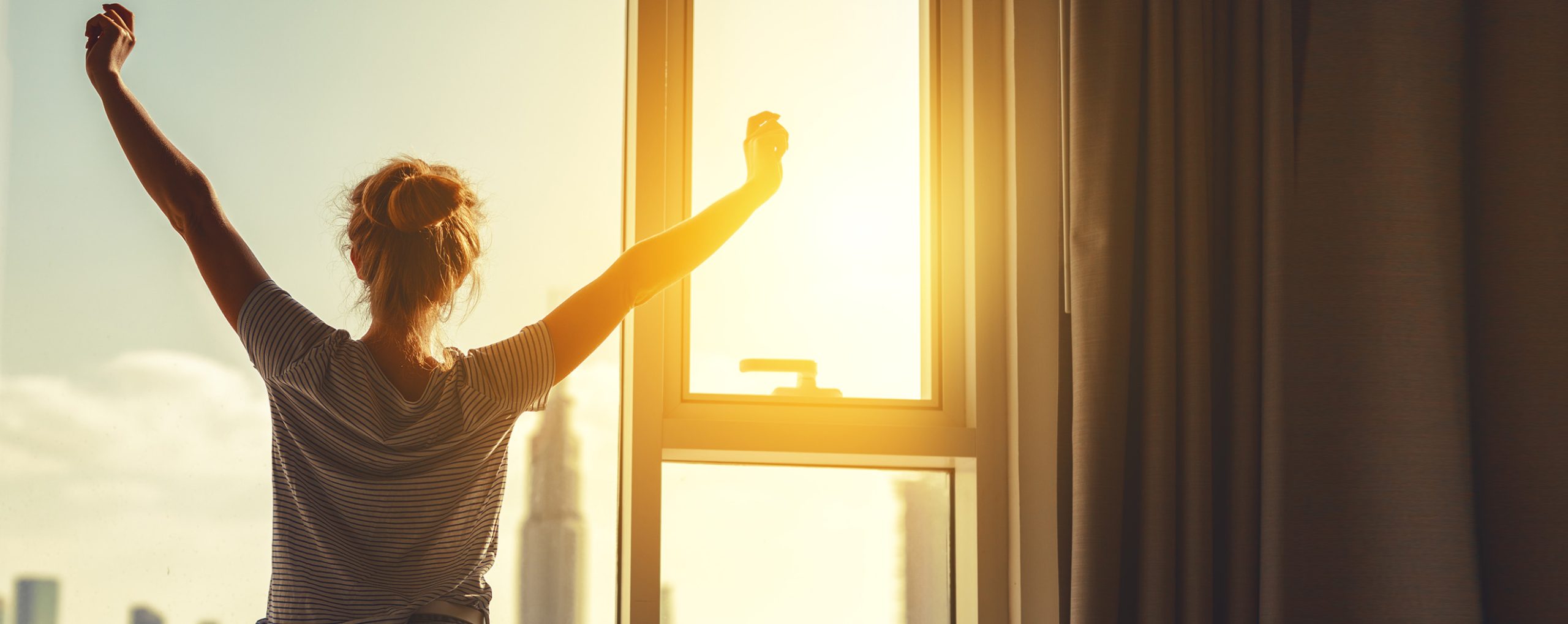 Frau streckt sich morgens vor einem Fenster, während die Sonne aufgeht.