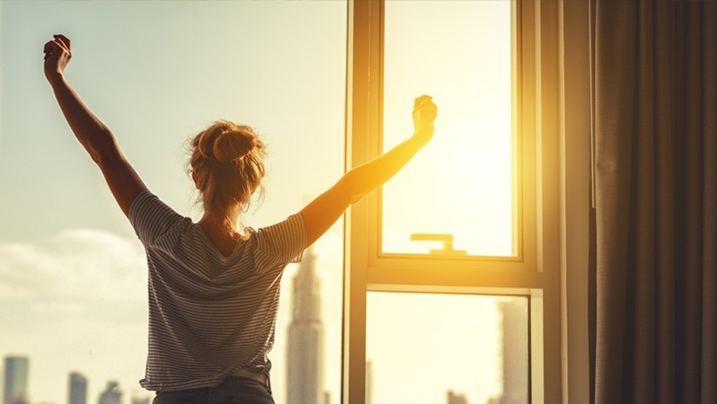 Frau streckt sich morgens vor einem Fenster, während die Sonne aufgeht.