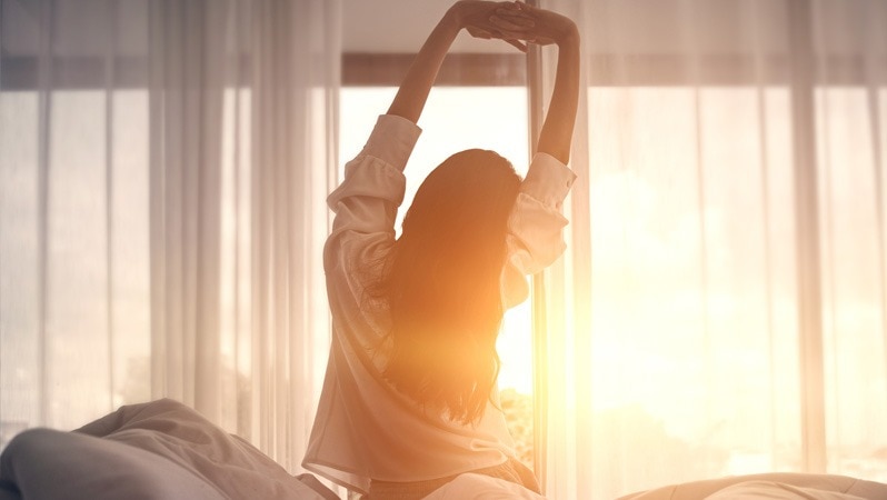 Frau streckt sich morgens vor einem Fenster mit Sonnenaufgang.