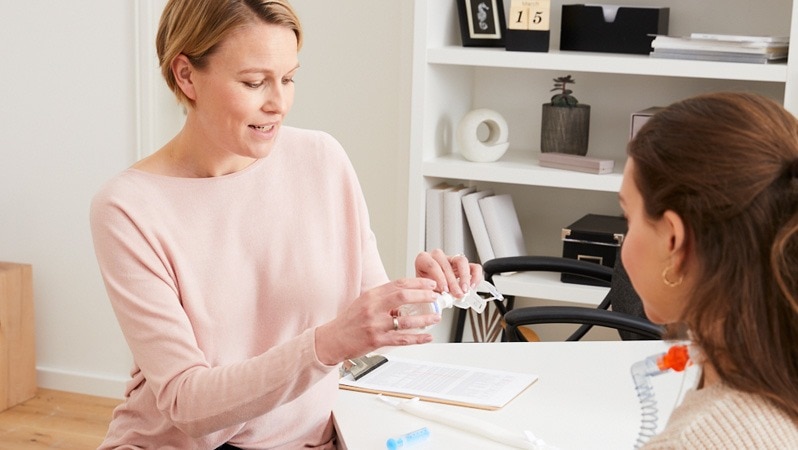 Eine medizinische Fachkraft erklärt einer Patientin die Handhabung einer Trachealkanüle. Beide sitzen in einem modernen Büro, während die Fachkraft detaillierte Anweisungen gibt.