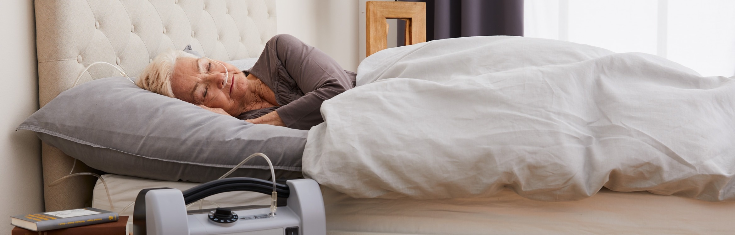 Eine ältere Frau mit Sauerstoffschlauch schläft friedlich in einem Bett, während ein Sauerstoffgerät neben dem Bett steht.