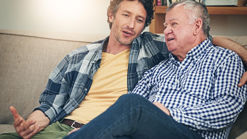 Ein älterer Mann sitzt entspannt auf einem Sofa, während ein jüngerer Mann neben ihm sitzt und ihm aufmerksam zuhört.