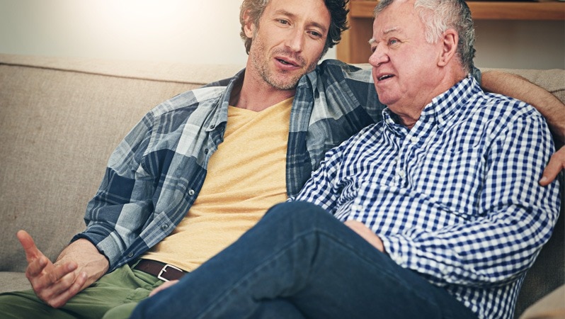 Ein älterer Mann sitzt entspannt auf einem Sofa, während ein jüngerer Mann neben ihm sitzt und ihm aufmerksam zuhört.