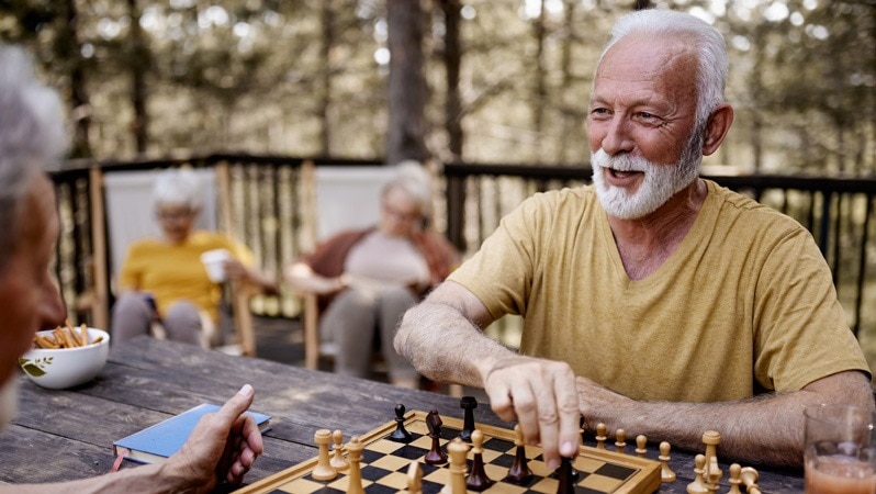 Ein älterer Mann spielt Schach auf einer Terrasse im Freien, während im Hintergrund weitere ältere Personen entspannt auf Stühlen sitzen.